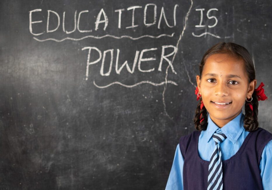 Smiling young girl kid with school uniform looking camera after writing education is power on black board at class room - concept of development, growth and wisdom.